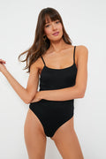 Woman in a black crinkled ribbed bodysuit with thin straps by onia, looking at the camera with one arm bent and the other relaxed.