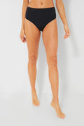A person stands barefoot wearing Solid & Striped high-waisted black ribbed bikini bottoms with a retro-inspired design.