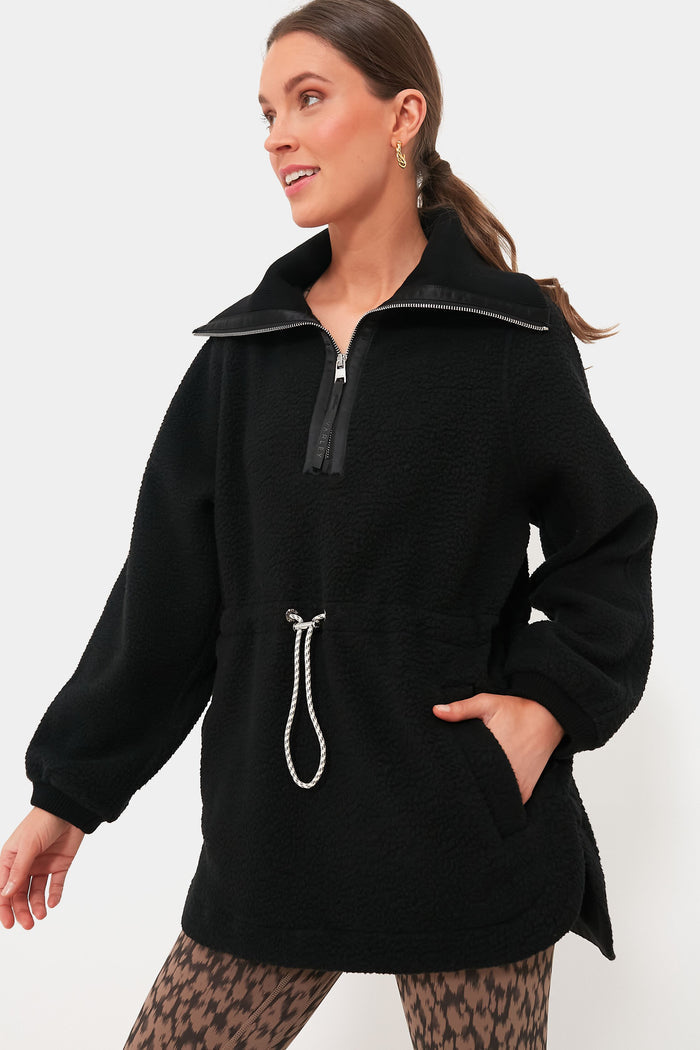 A woman wears a Varley black plush Borg fleece half-zip pullover with a drawstring waist, Alpine loungewear patterned leggings, and gold earrings.