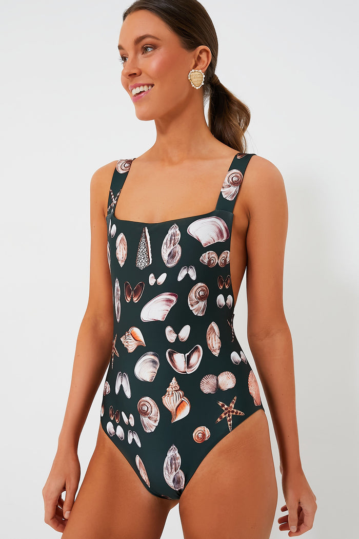 A woman smiles warmly wearing AGUA BY AGUA BENDITA’s black one-piece swimsuit featuring a seashell pattern, crafted from recycled polyester.