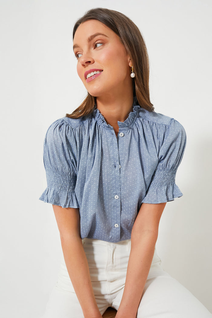 A woman seated and looking to her left wears the Mille Chambray Polka Dot Marnie Top featuring short puff sleeves and a ruffle neckline, paired with white pants.