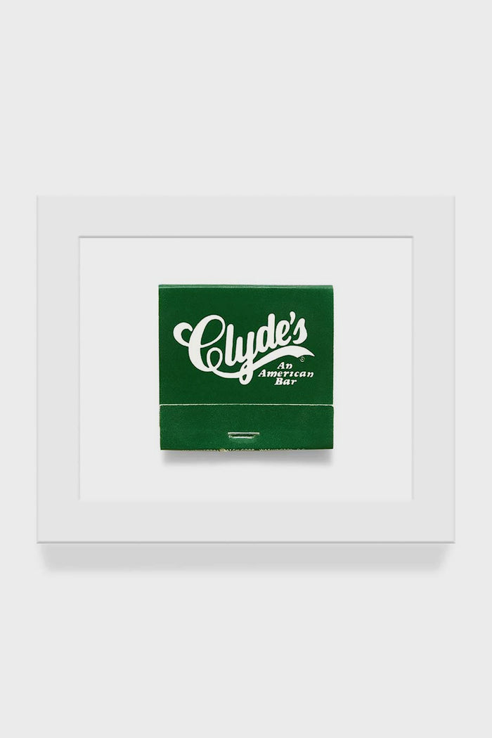 A green matchbook with "Clydes An American Bar" is centered in a white frame, resembling framed art. From Matchbook Diaries by Charles Ryan Clarke.