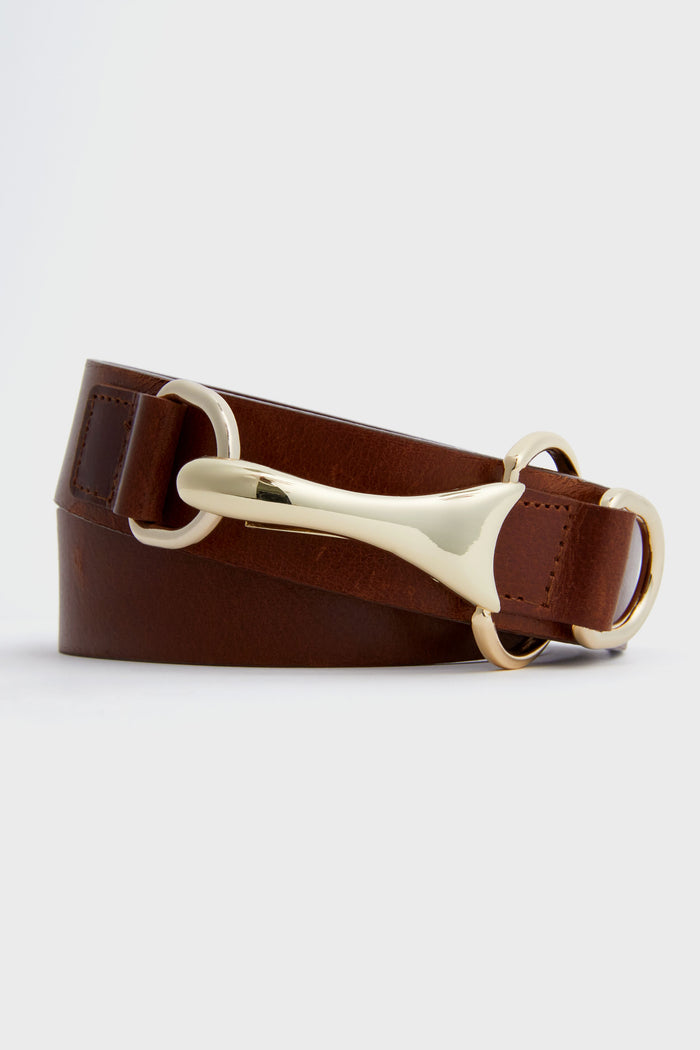 A Tuckernuck Accessories Cognac Snaffle Belt in brown with gold-tone hardware, featuring a large decorative gold buckle and circular rings, is shown coiled.