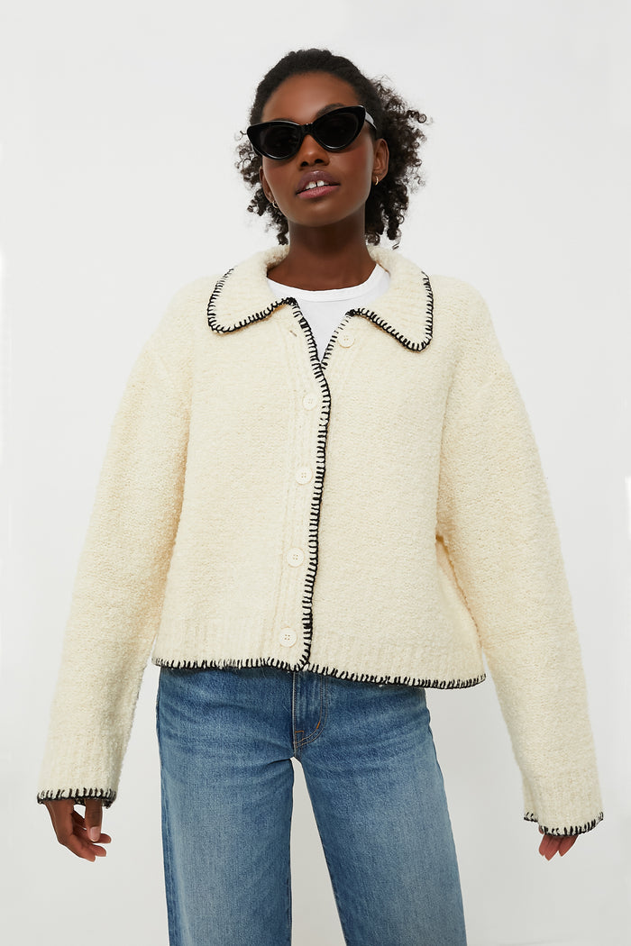 A person wearing black sunglasses, a cream Moon River cardigan with contrast stitching and black trim, and blue jeans stands confidently.