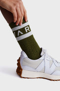 A hand adjusts an olive green Varley sock with white lettering above a white sneaker with a gum-textured sole.