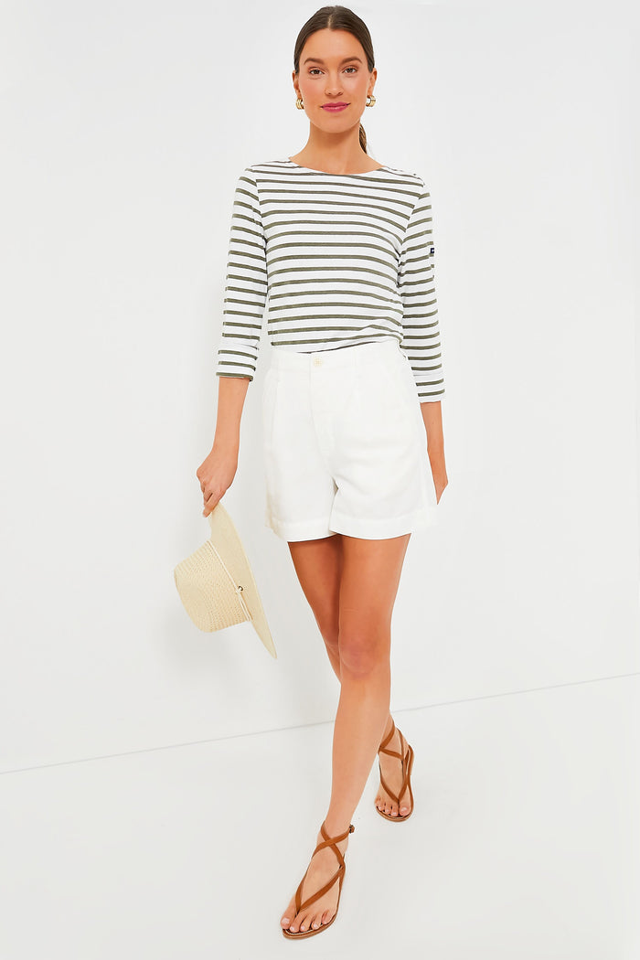 A woman wears a striped long-sleeve top, brown sandals, and holds a straw hat paired with Alex Mill Ecru Twill Pleated Shorts—the perfect warm-weather staple.