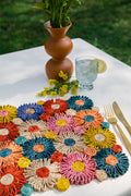 A multicolored handmade placemat by Payton James on a white table with a glass of water, gold cutlery, and a brown vase of flowers.