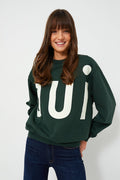 A woman with long brown hair wears a dark green Clare V Oui print sweater and dark blue jeans, with "Oui" in large white letters.