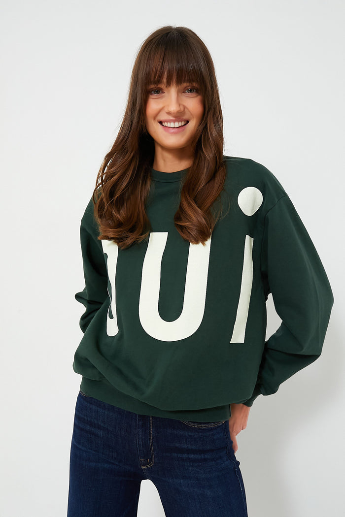 A woman with long brown hair wears a dark green Clare V Oui print sweater and dark blue jeans, with "Oui" in large white letters.