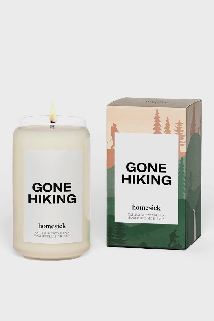A lit pine-scented soy wax candle labeled GONE HIKING sits beside its illustrated box, both featuring Homesick Candles branding below the label, bringing a cozy nature-inspired fragrance to your home.