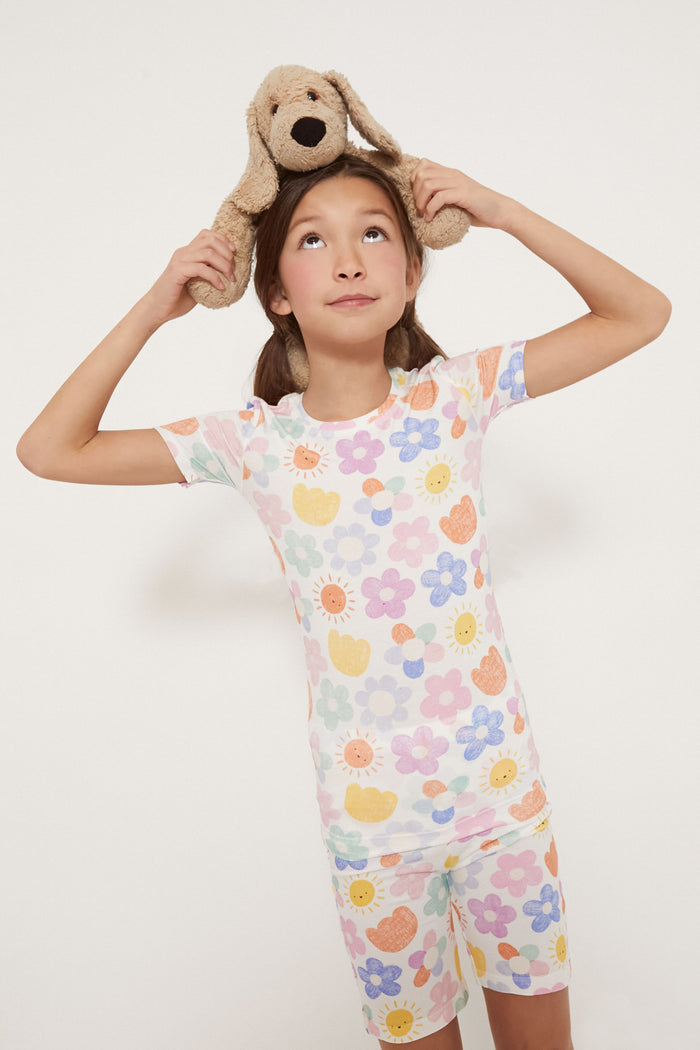 A young girl wearing Angel Dear bamboo baby pajamas with floral prints holds a stuffed dog on her head and looks upward.