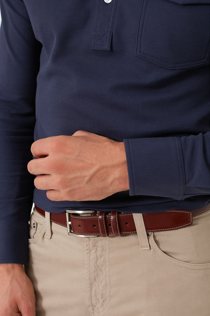 A person in a navy long-sleeve shirt, tan pants, and a Trafalgar Dropped Edge full grain leather belt with silver buckle demonstrates how luxury men’s accessories can elevate any outfit. Their left hand rests casually on their waist.