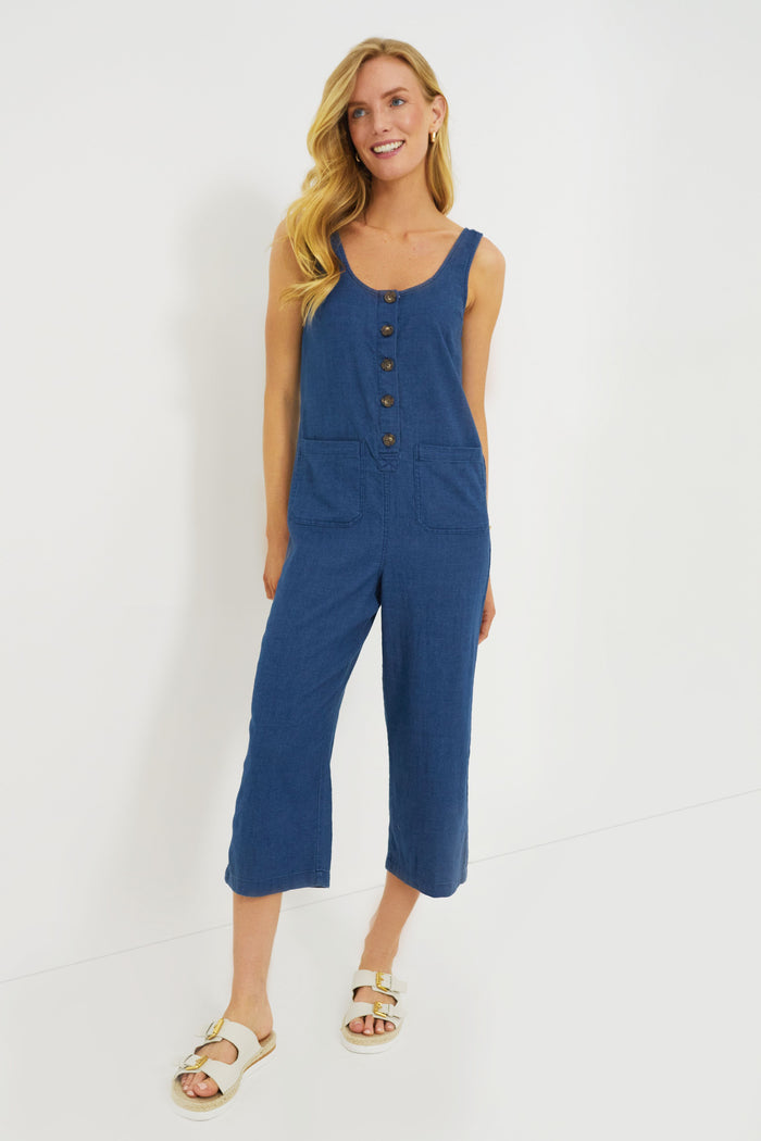 A woman wears a sleeveless blue Marine Layer cotton jumpsuit with two front pockets and beige sandals.
