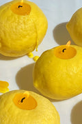 Three yellow lemons are hollowed out and filled with soy wax and wicks, creating unique handmade lemon candles by Scandles that burn brightly on a white surface.