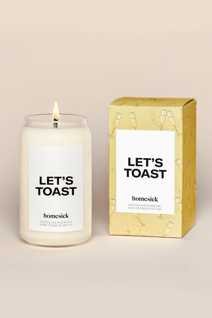 A Homesick Candles Let's Toast candle glows in a glass jar labeled LET'S TOAST, next to its yellow box with champagne illustrations, bringing a festive vibe to your home fragrance collection.