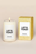 A Homesick Candles Let's Toast candle glows in a glass jar labeled LET'S TOAST, next to its yellow box with champagne illustrations, bringing a festive vibe to your home fragrance collection.