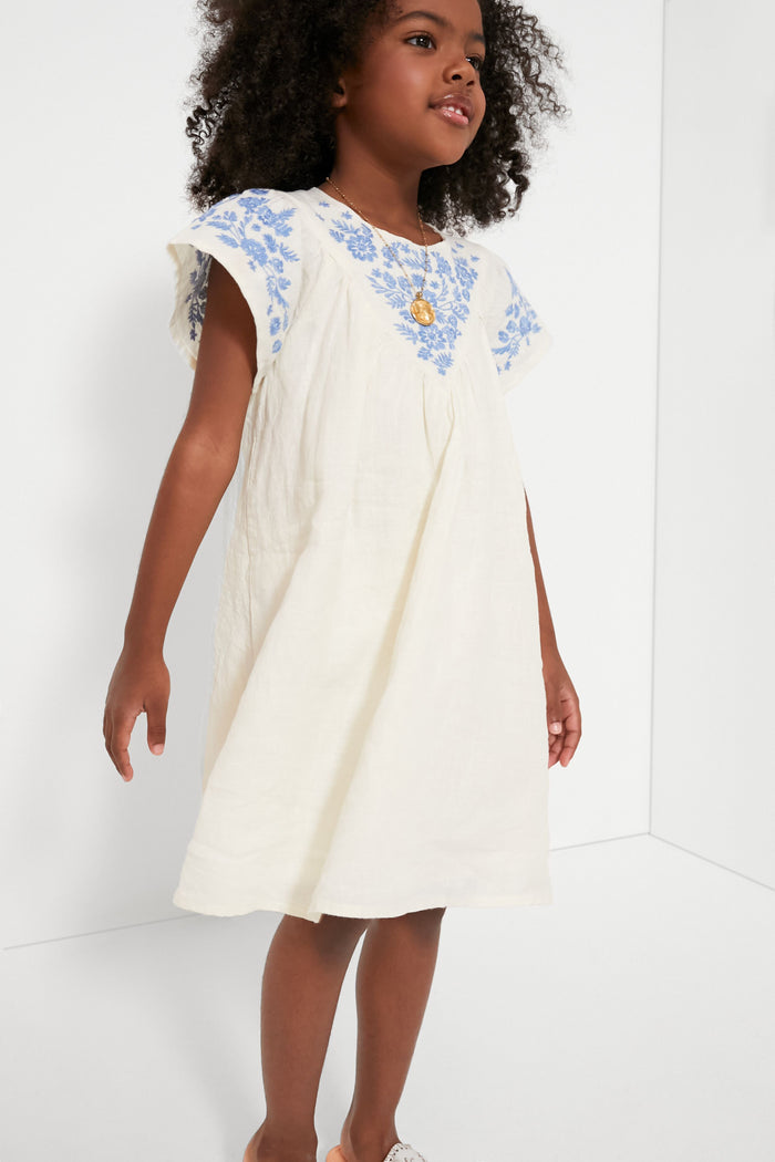 A young girl wears a Tartine et Chocolat white linen dress with blue floral embroidery on the yoke, short flutter sleeves, and a gold necklace—a charming example of French childrenswear.