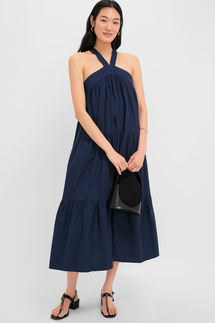 A woman in the En Saison Navy Irene Dress, featuring an A-line silhouette and halter neckline, pairs it with black sandals and a black bag while standing against a plain light background.