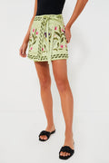 A woman wears an AGUA BY AGUA BENDITA light green embroidered mini pareo wrap skirt with a tie belt, styled with a black top and black slide sandals for a chic, relaxed look.
