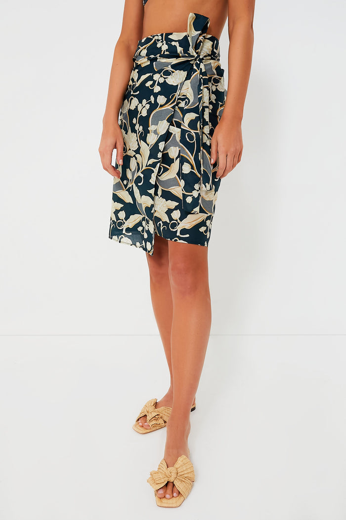 A woman wears an AGUA BY AGUA BENDITA navy blue and beige floral mini cover-up wrap skirt with a side tie, crafted from a cotton-silk blend, paired with beige woven sandals.
