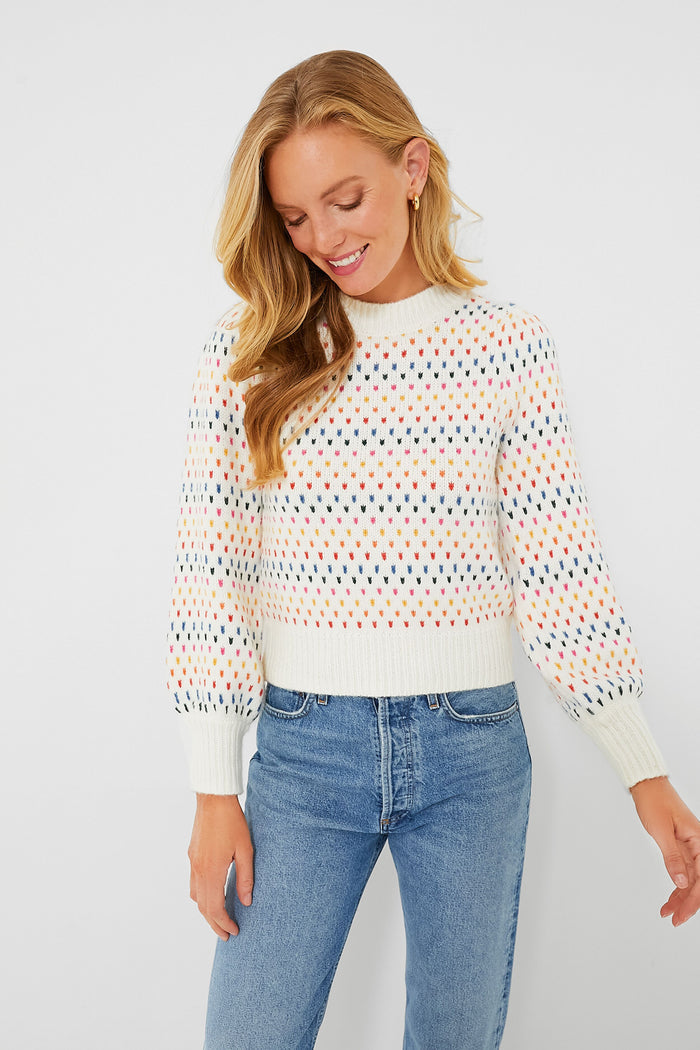 A woman in a Marine Layer white puff sleeve sweater with a multicolored dot pattern and blue jeans looks down with a slight smile.