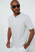 A man in black sunglasses, white pants, and a Faherty short-sleeve white and gray striped organic cotton henley showcases rugged style with sustainable apparel.