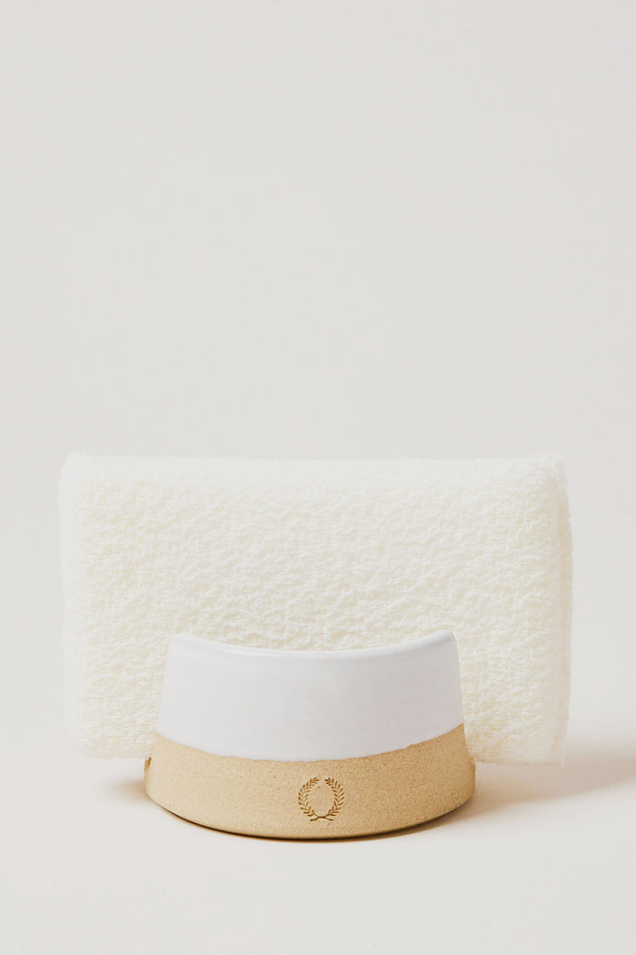 A white textured towel stands upright behind a beige and white hat with a laurel wreath emblem, alongside Farmhouse Pottery, Inc accents and a kitchen sponge holder.