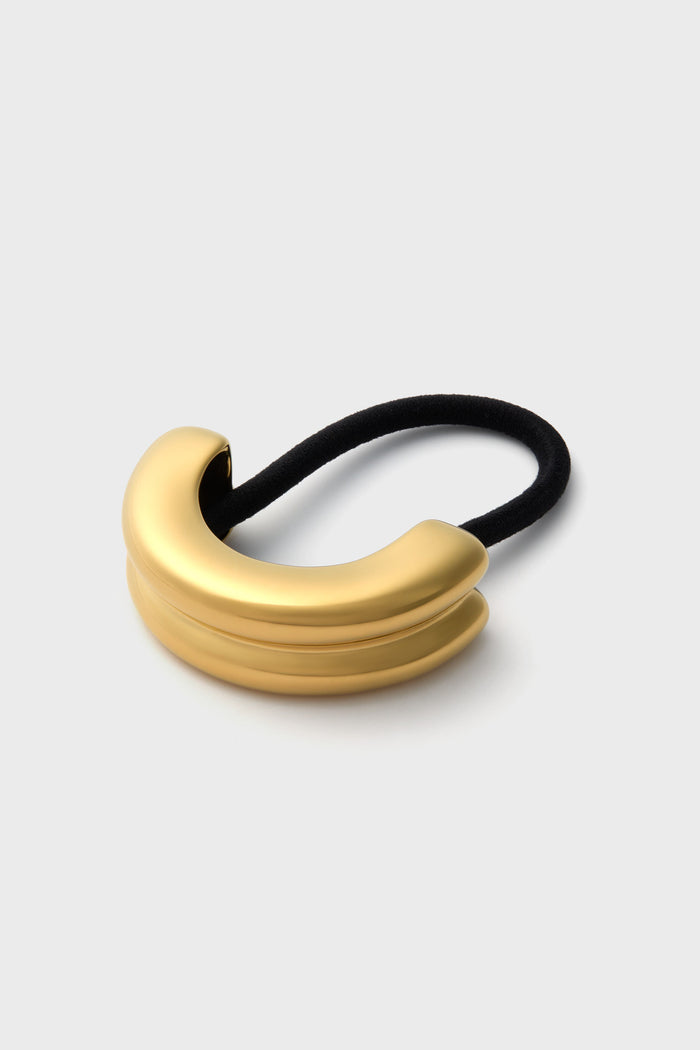 A gold-plated curved metal hair tie by Petit Moments features a black elastic band, making it an elegant hair accessory.