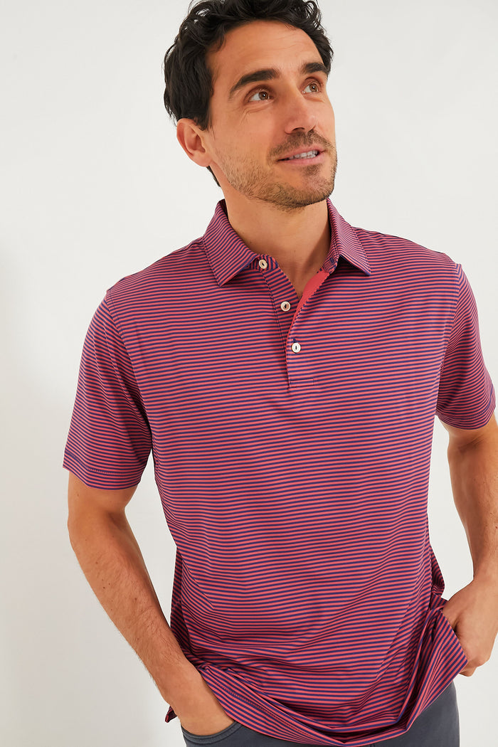 A man wears a Peter Millar short-sleeved red and blue striped performance jersey polo with UPF 50+ protection, looking up with hands in his pockets.