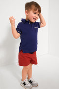 A young boy beams with arms raised, dressed in Oso & Me durable children’s wear: a navy polo shirt, adjustable waist red shorts, and gray sneakers with white and black accents.