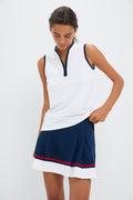 A woman wearing a Tnuck Sport sleeveless mock neck white top with a zippered collar and a navy pleated skirt with red and white stripes looks down, holding the hem of her top.